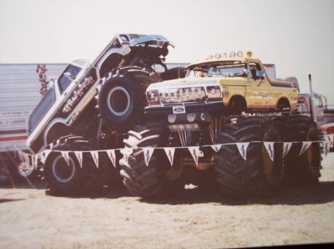 1985 Ford Bronco - Two monster trucks are positioned on display, one atop the other, with large tires and custom paint jobs.