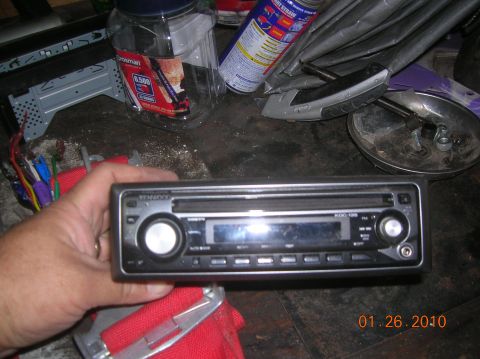 1985 Ford Bronco - A Kenwood car stereo is held in a hand, with various other items scattered around the surface.