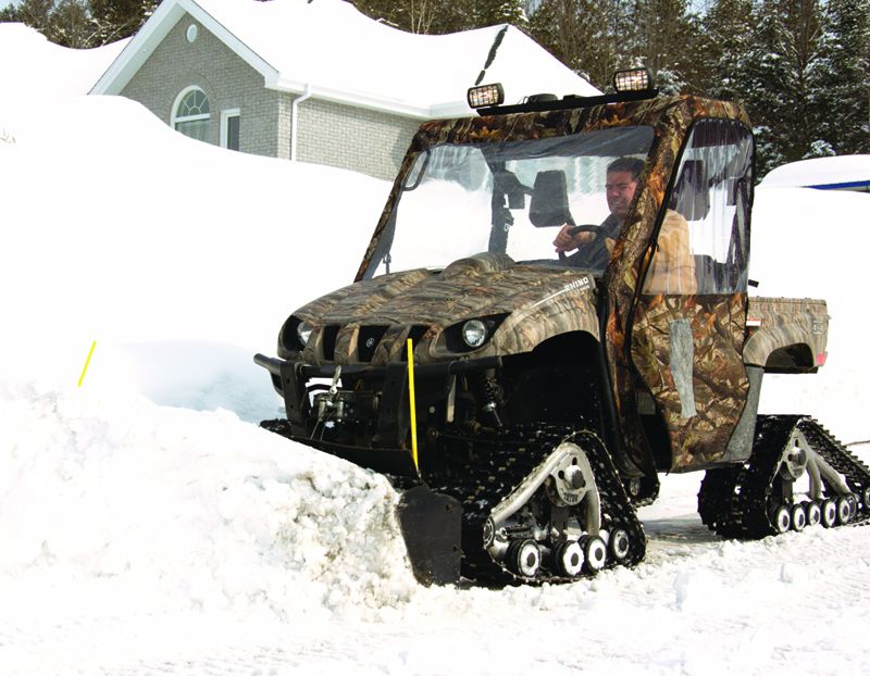 Camo UTV on rubber tracks driving through deep snow near a house