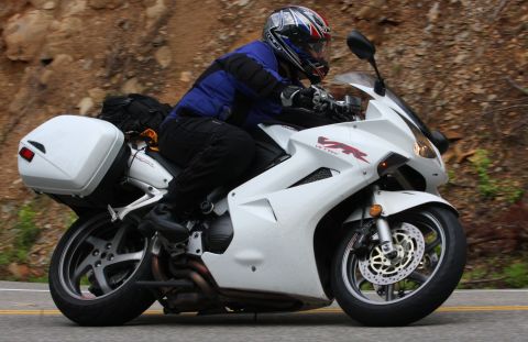 1985 Ford Crown Victoria LTD - A rider is on a white Honda VFR motorcycle equipped with saddlebags, traveling on a road with a rocky embankment.