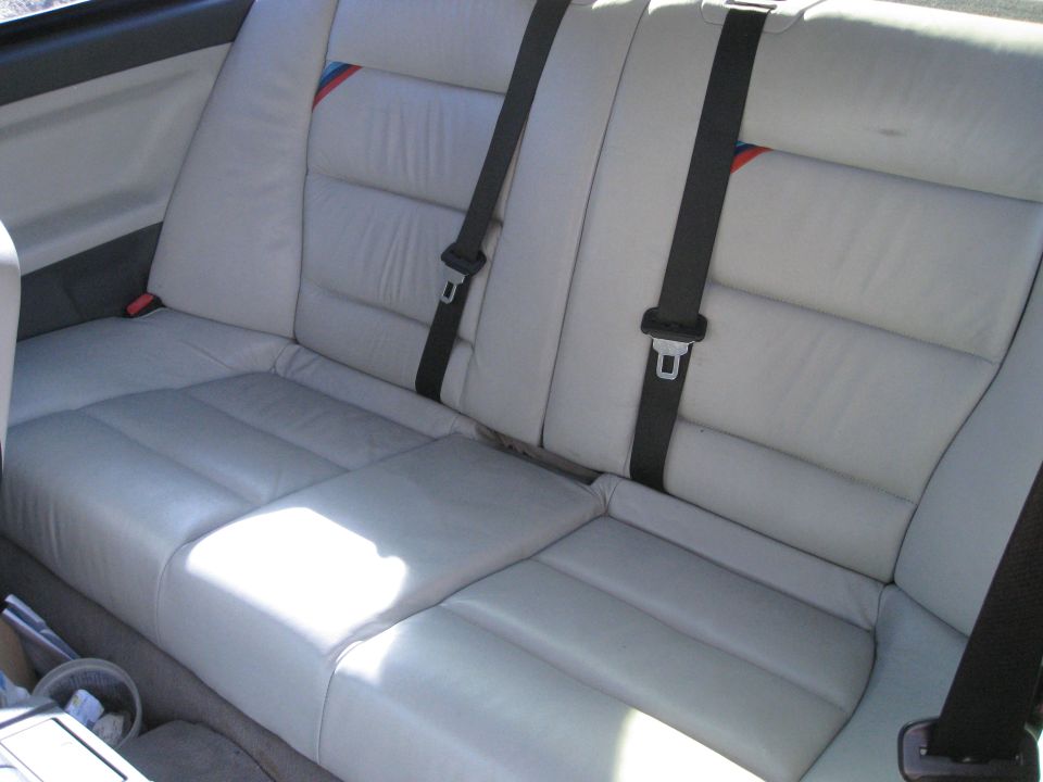 Light gray leather rear car seats with black seatbelts and a red, white, and blue stripe detail on the headrest