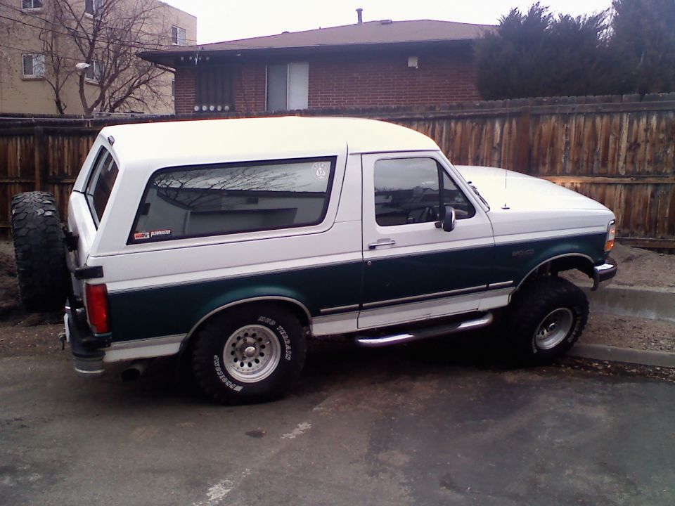 White and green Ford Bronco parked on pavement, spare tire mounted on rear. Lifted suspension and off-road tires visible