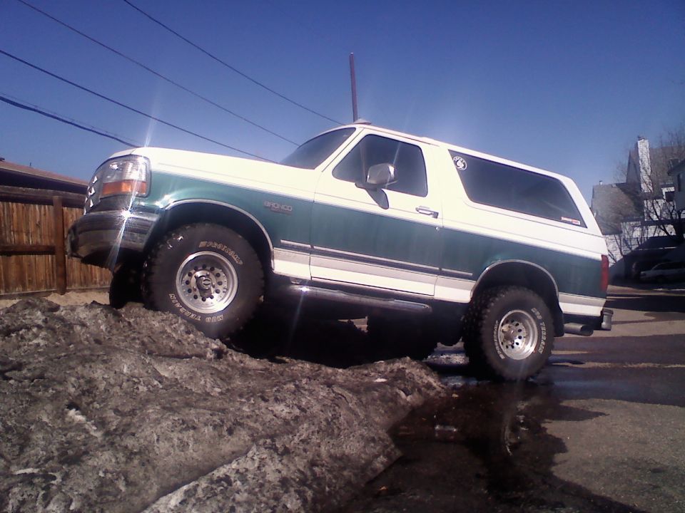 Green and white lifted Ford Bronco on mud-terrain tires climbing a snowbank, driver's side view