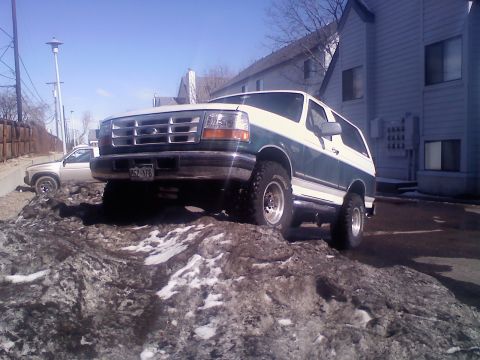 1996 Ford Bronco - Low three-quarter front view of a lifted teal-green and white two-tone Ford Bronco climbing a plowed pile of dirty snow and mud beside a gr…