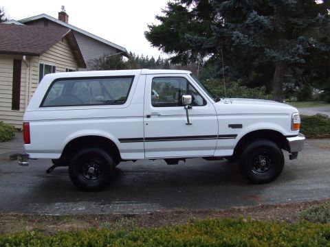 1996 Ford Bronco - White 1996 Ford Bronco parked on a paved surface in front of a house and trees.