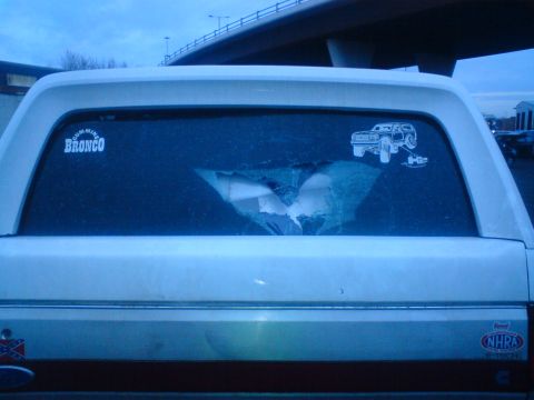 1989 Ford Bronco - Rear view of a white SUV with a broken window and various stickers, including one for Bronco.