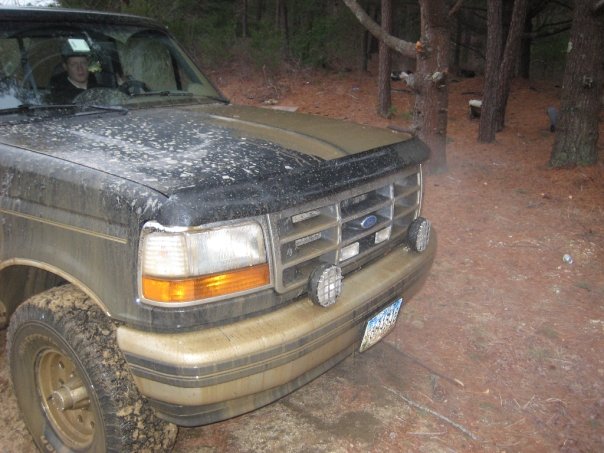 Front end of a dirty Ford Bronco parked in a wooded area. A person is visible in the driver's seat