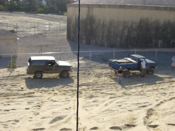 Brown Bronco and blue pickup truck on sandy terrain near a large concrete structure