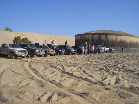 1994 Ford Bronco - Several off-road vehicles are gathered on sandy terrain near a large, round concrete structure under a clear blue sky.
