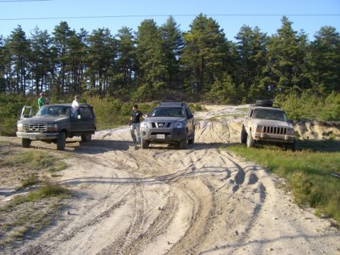 1994 Ford Bronco - Multiple SUVs navigate a challenging dirt path surrounded by pine trees under a clear blue sky.
