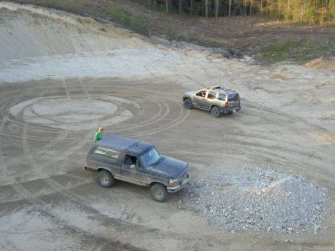 1994 Ford Bronco - Two SUVs are seen in a dirt lot, one dark blue Bronco and a silver SUV.
