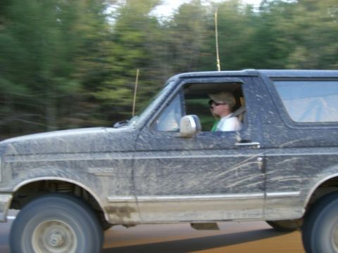 1994 Ford Bronco - A driver navigates a muddy 1994 Ford Bronco down a road, with trees blurred in the background.