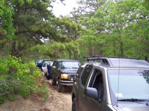 1994 Ford Bronco - Several SUVs are driving in a convoy on a dirt path surrounded by trees.