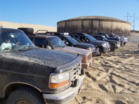 1994 Ford Bronco - A line of off-road vehicles are parked on a sandy terrain under a clear blue sky.