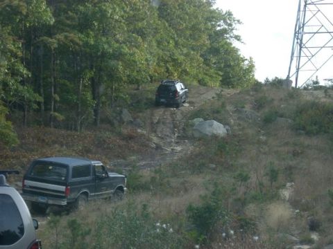 1994 Ford Bronco - Two SUVs navigate a rocky, wooded incline near a large transmission tower.