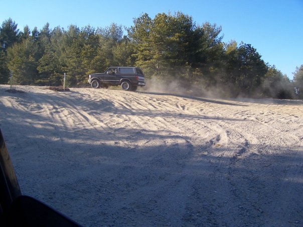 1994 Ford Bronco driving on a dirt road, kicking up dust. Trees and blue sky in background