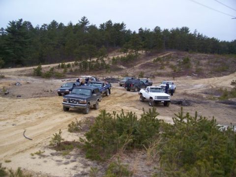 1994 Ford Bronco - A group of off-road vehicles are gathered in a sandy, wooded area on an overcast day.