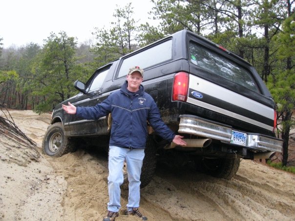 Black Ford Bronco with a man standing in front, angled rear view, on a sandy incline