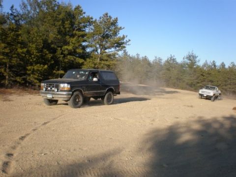 1994 Ford Bronco - A black Ford Bronco drives on a dirt road with a white truck in the distance.