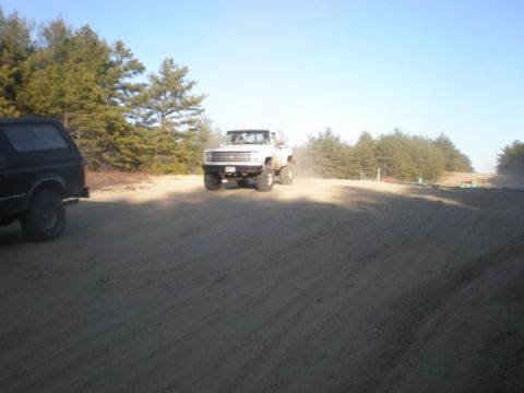 1994 Ford Bronco - A white pickup truck kicks up dust as it drives on a dirt road, with a black SUV visible on the left.