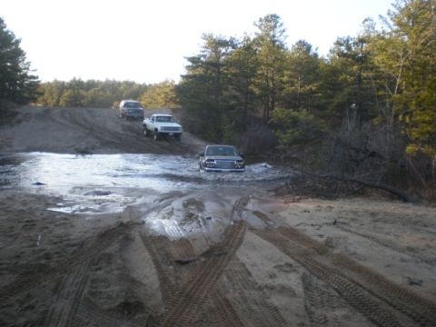 1994 Ford Bronco - Several trucks navigate a challenging off-road course with mud and water obstacles.