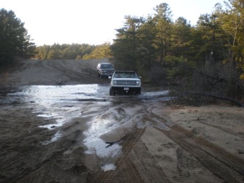 1994 Ford Bronco - Two SUVs navigate a muddy, water-filled track surrounded by trees.