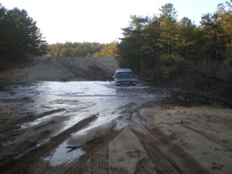 1994 Ford Bronco - A dark SUV navigates through a water-filled dirt road, surrounded by trees and sandy terrain.