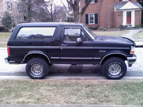 1994 Ford Bronco - This black Ford Bronco is parked on the side of the road in front of a brick house.