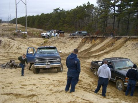 1994 Ford Bronco - Several trucks and SUVs are gathered in a sandy, off-road area with trees in the background.
