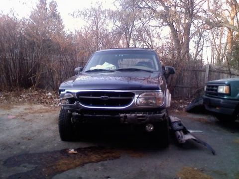2000 Ford Explorer - A dark blue Ford Explorer sits in a yard with front-end damage, next to another vehicle.