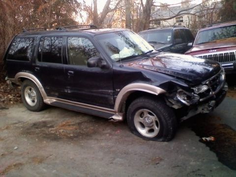 2000 Ford Explorer - A damaged dark SUV sits parked next to other vehicles, showing a flat tire and bent bodywork.