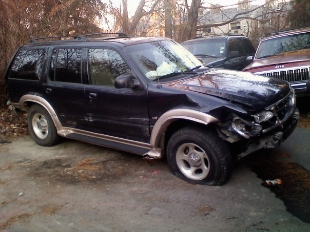 Dark SUV with a flat front tire and crumpled front fender, seen from the front-left