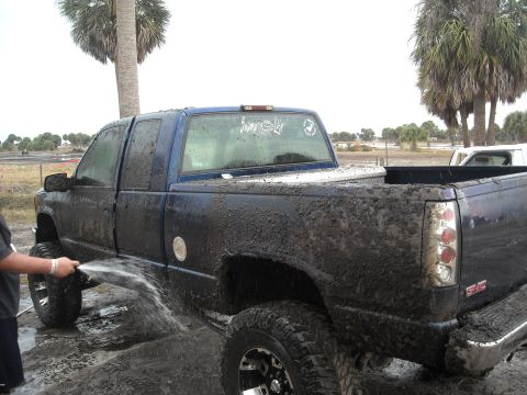 1992 Ford Bronco - A person washes a heavily mud-covered GMC pickup truck in an outdoor setting with palm trees.