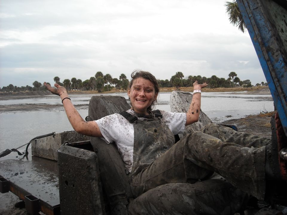 Mud-covered person in overalls sits in a vehicle seat, arms outstretched