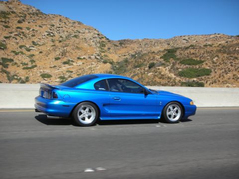 1998 Ford Crown Victoria - A bright blue coupe is captured in profile while driving on a highway, with a dry, rocky hillside under a clear blue sky.