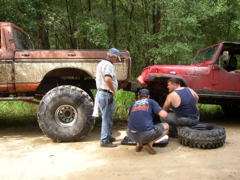 1991 Ford Bronco - Two men appear to be fixing a red Jeep while a large, lifted truck sits nearby in a wooded area.