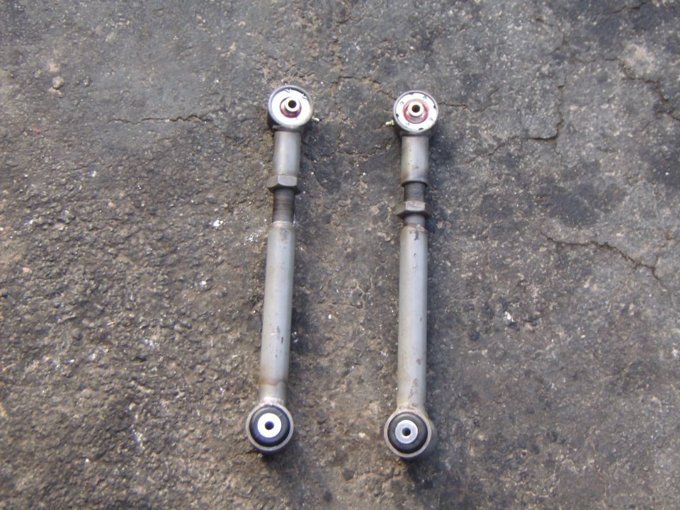 Two silver adjustable control arms with rubber bushings on a concrete surface. Supermotors.net logo in the bottom right
