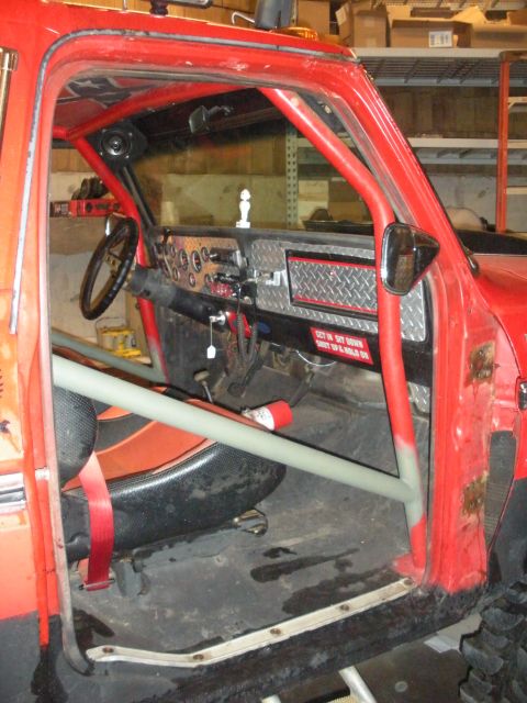 1979 Ford Bronco - Driver's-door opening of a red Ford Bronco trail truck with a gray tubular roll cage running through the cab and a door bar across the open…