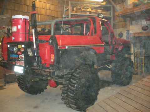 1979 Ford Bronco - Red early Ford Bronco built as a trail rig, parked inside a wood-plank barn and seen from the rear three-quarter. It has an exposed tubular…