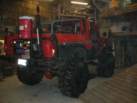1979 Ford Bronco - A red lifted Ford Bronco viewed from the rear three-quarter angle, parked inside a wood-walled garage. It features an exposed roll cage, la…