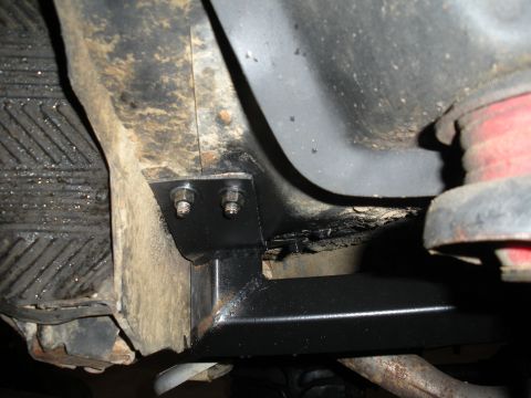 1979 Ford Bronco - Close-up under a vehicle showing a black-painted fabricated bracket bolted to the frame rail with two hex bolts, tying into a welded tube o…