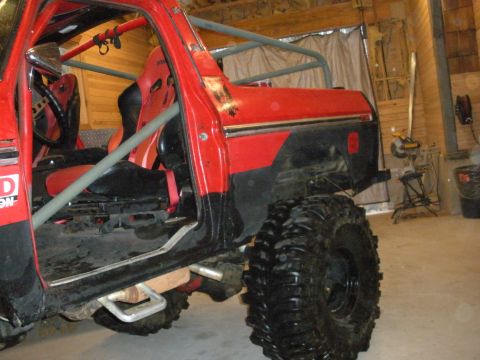 1979 Ford Bronco - A red 1970s-era Ford Bronco SUV shown from the side with the doors removed, revealing a gray tubular roll cage, red racing seat, and steeri…