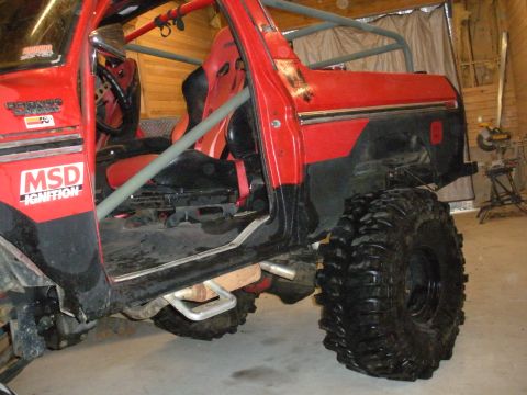 1979 Ford Bronco - Low side view of a doorless red Ford Bronco in a wood-lined garage, showing the gray tube roll cage running through the cab and a red racin…