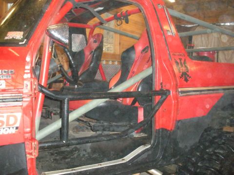 1979 Ford Bronco - Side view into the stripped cab of a doorless red Ford Bronco showing a gray tube roll cage with door-bar tubes, two red and black racing b…