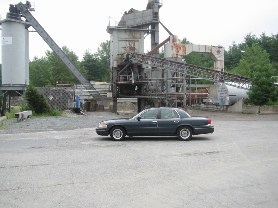 Dark green Mercury Grand Marquis sedan parked on pavement in front of an industrial asphalt plant