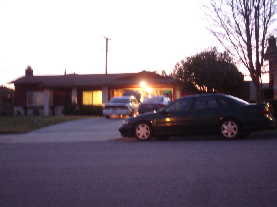 Dark sedan parked on a driveway, seen from the side at dusk. Other cars are visible in the background