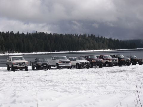 1991 Ford Bronco - A fleet of SUVs and trucks, including a white 1991 Ford Bronco, are parked in a snowy landscape next to a body of water.