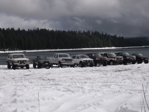 1991 Ford Bronco - Several SUVs and trucks are parked in a line on a snowy landscape next to a body of water.