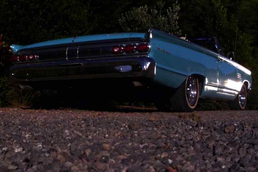 Rear view of a teal 1965 Mercury Park Lane convertible parked on a gravel surface with trees in the background