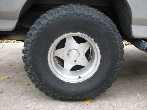 Close-up of a five-star alloy wheel with a knobby off-road tire. Part of a silver vehicle body is visible above